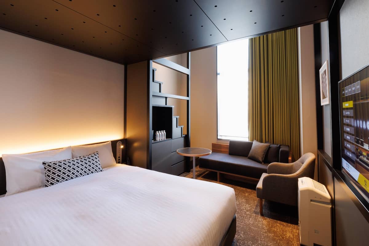 A modern hotel room with a large bed, a seating area with a small table, and a TV screen displaying a menu.