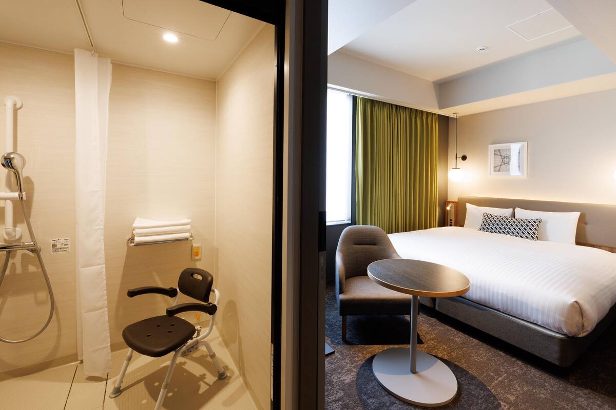 A hotel room with a bed, a small round table, a chair, and a shower area.
