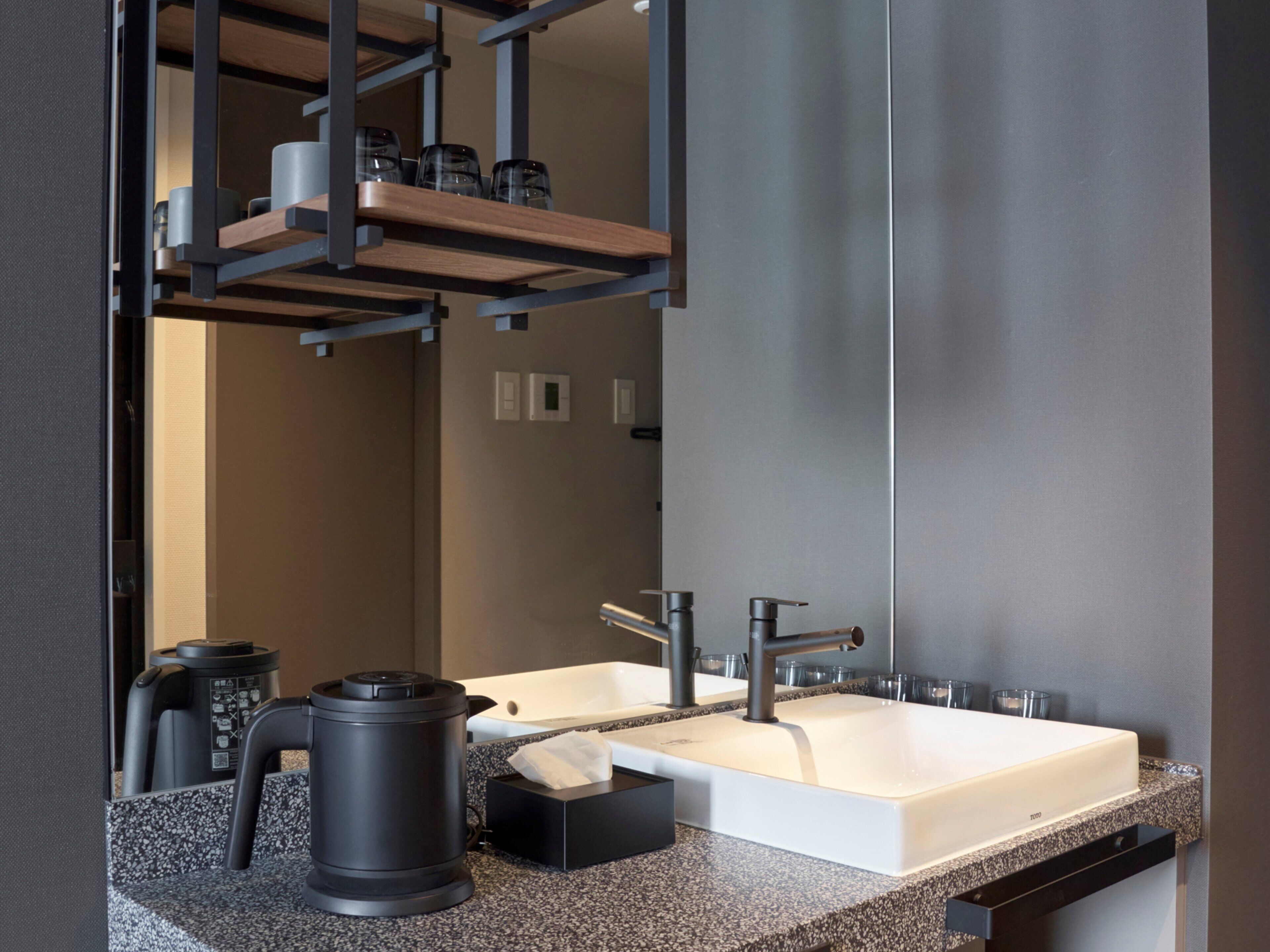 A modern bathroom with a large mirror, two sinks, a coffee machine, and a trash bin.