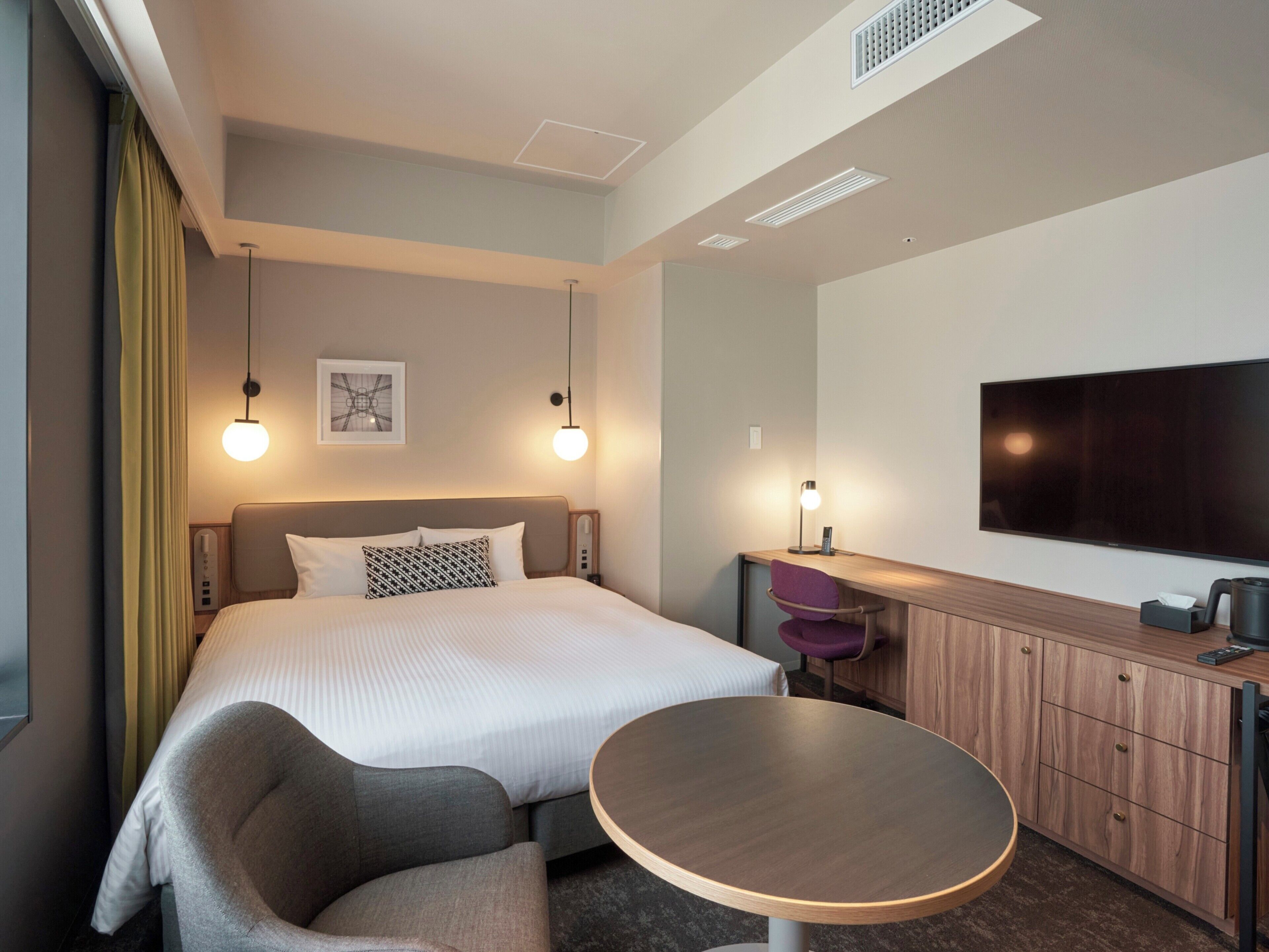 A modern hotel room with a bed, a desk, a chair, a round table, a TV, and a ceiling light.