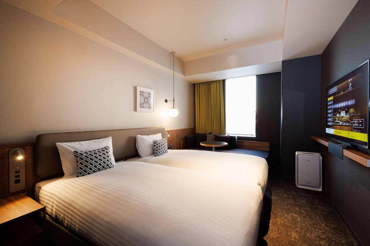 A hotel room with a large bed, bedside tables, a small round table, a TV, and a window with curtains.