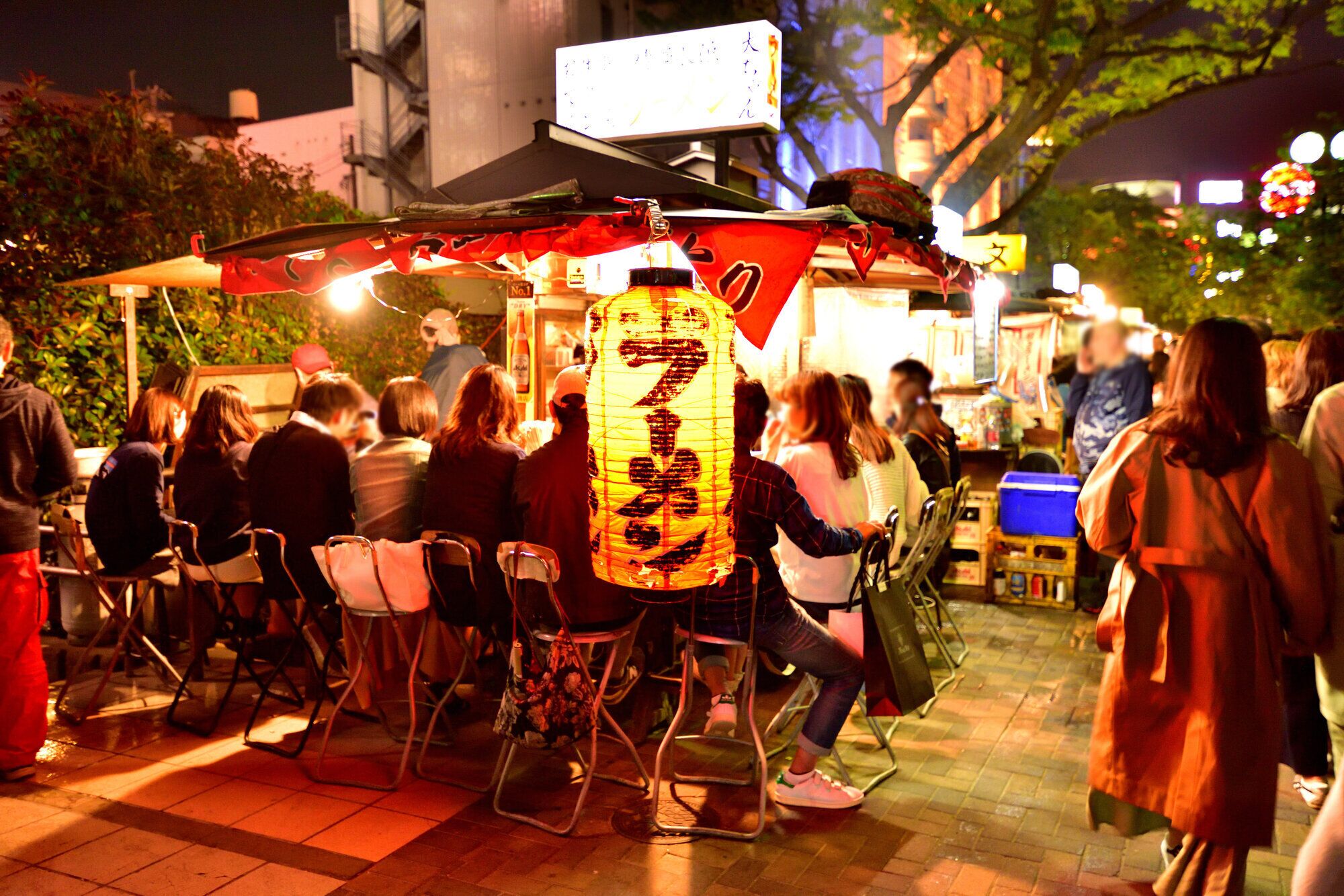 A street food stall with a large illuminated sign, outdoor seating, and a crowd of people.
