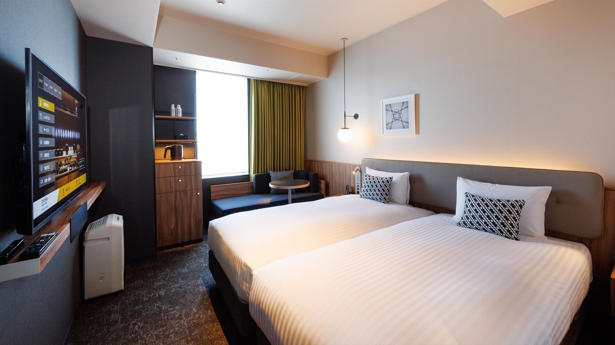 A hotel room with a bed, a desk with a computer, a small table, a sofa, and a window with curtains.