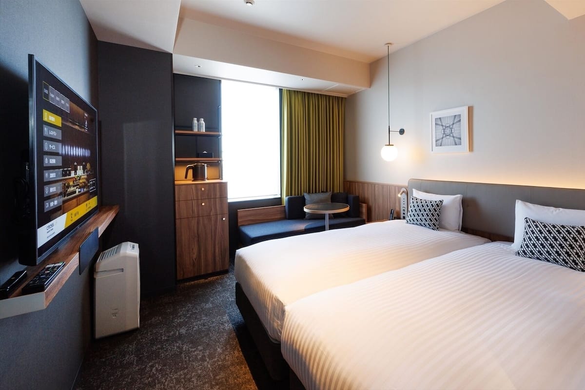 A hotel room with a bed, a desk with a computer, a small table, a sofa, and a window with curtains.