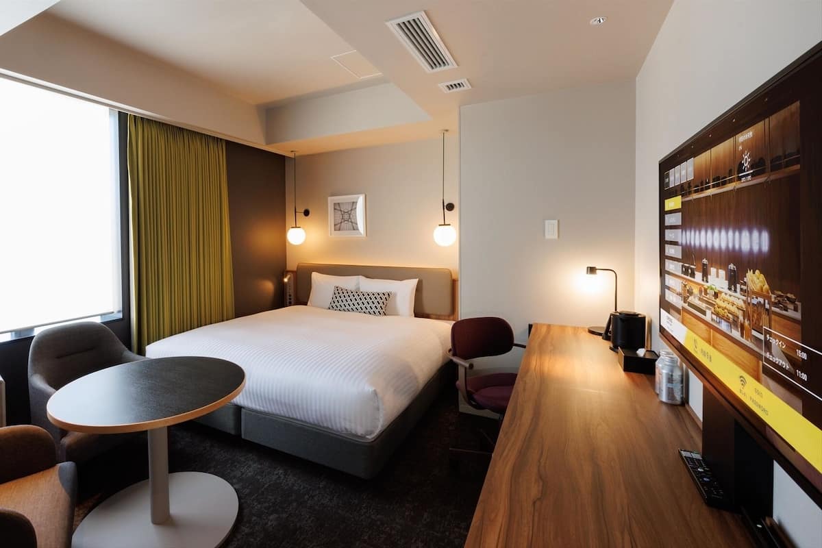 A hotel room with a bed, a desk, a chair, a small table, a TV, and a window with curtains.