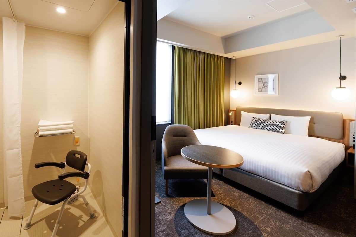 A hotel room with a bed, a chair, a small table, a shower, and a window with curtains.