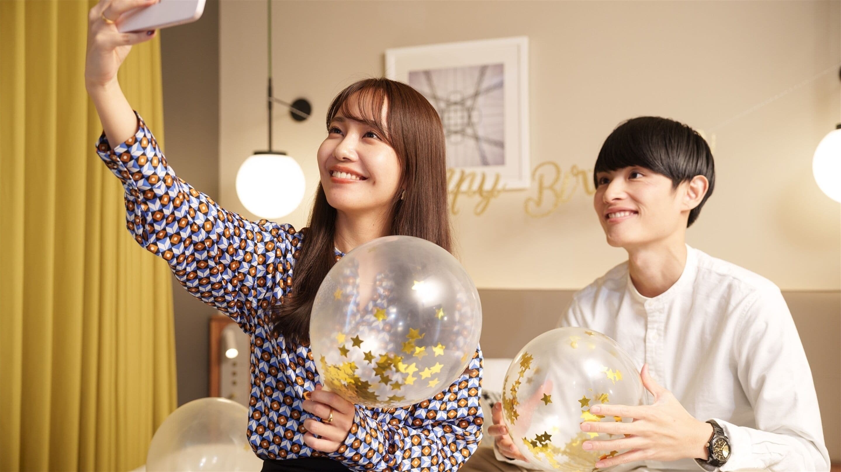 Two people holding transparent balloons with gold stars, taking a selfie in a room with a 'Happy Birthday' sign and a framed picture on the wall.