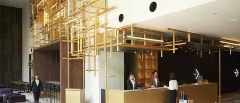 A reception area with a modern wooden reception desk, a seating area with purple chairs, and a reception desk with a computer and a person standing behind it.