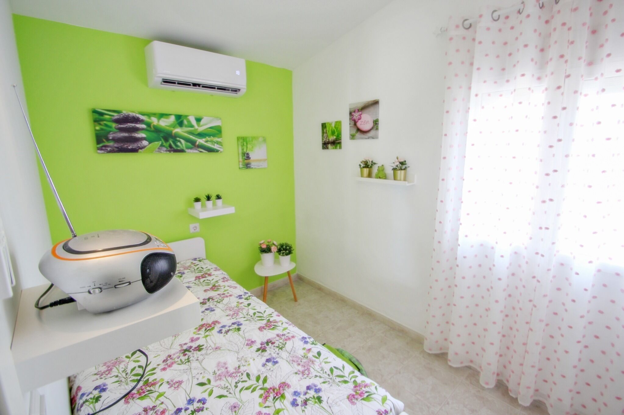 Una habitación con pared verde, una cama con ropa de cama floral, una mesita blanca con plantas y un aire acondicionado blanco.