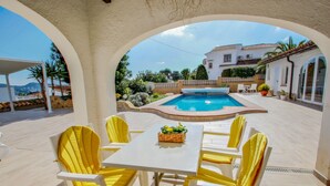 A poolside area with a table and chairs, a pool, and a view of a white house in the background. - La Tranquilidad - private pool villa with panoramic views in Moraira (Teluda)