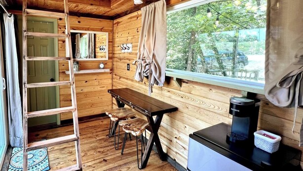 A wooden cabin interior with a wooden table, two chairs, a ladder, a window with curtains, and a coffee maker. - Table Rock Lake Tiny House Container Home Get Away (Shell Knob)
