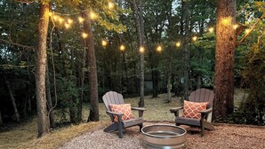 An outdoor seating area with Adirondack chairs and a metal fire pit surrounded by gravel, set in a forested area with string lights overhead. - Table Rock Lake Tiny House Container Home Get Away (Shell Knob)