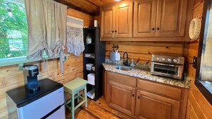 A compact kitchen with wooden cabinets, a built-in oven, a coffee maker, and a small stool. - Table Rock Lake Tiny House Container Home Get Away (Shell Knob)