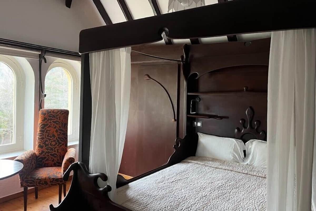 Een hemelbed met witte lakens, een gestreept fauteuil en uitzicht op het groen door boogvensters.