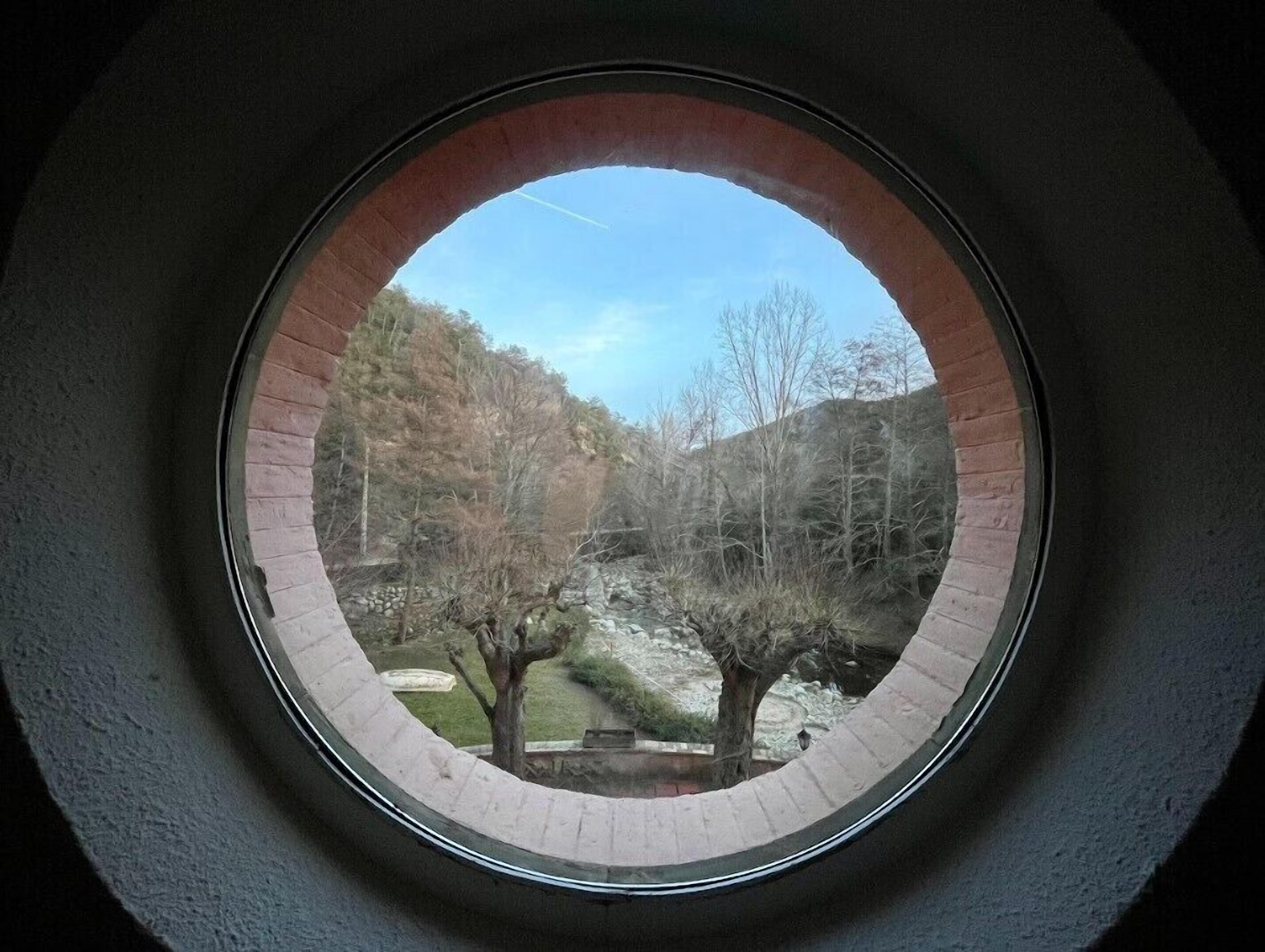 A circular window with a view of a natural landscape, including trees and a hill.