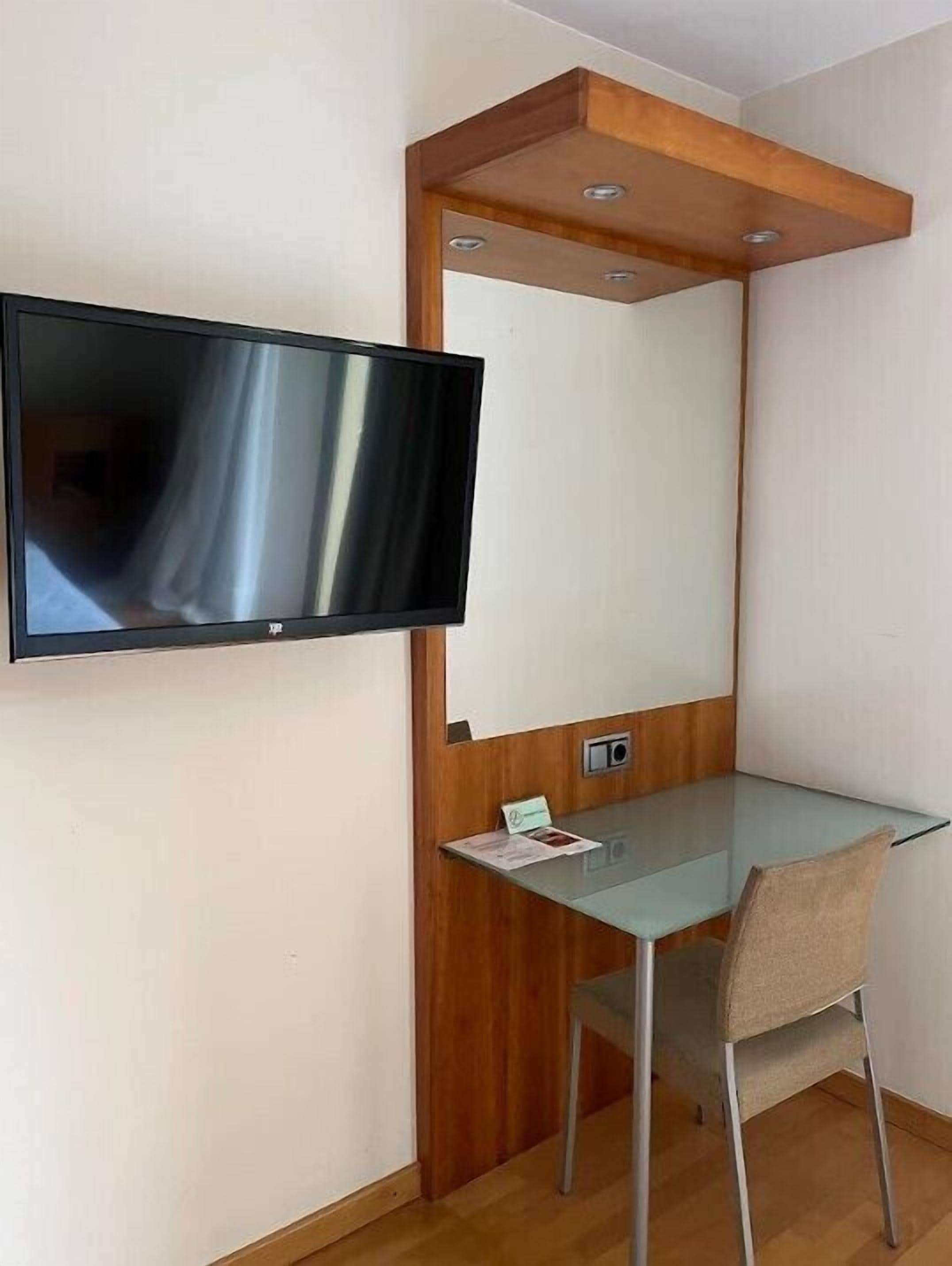 A small, modern room with a flat-screen TV mounted on the wall, a glass desk, and a wooden accent wall.