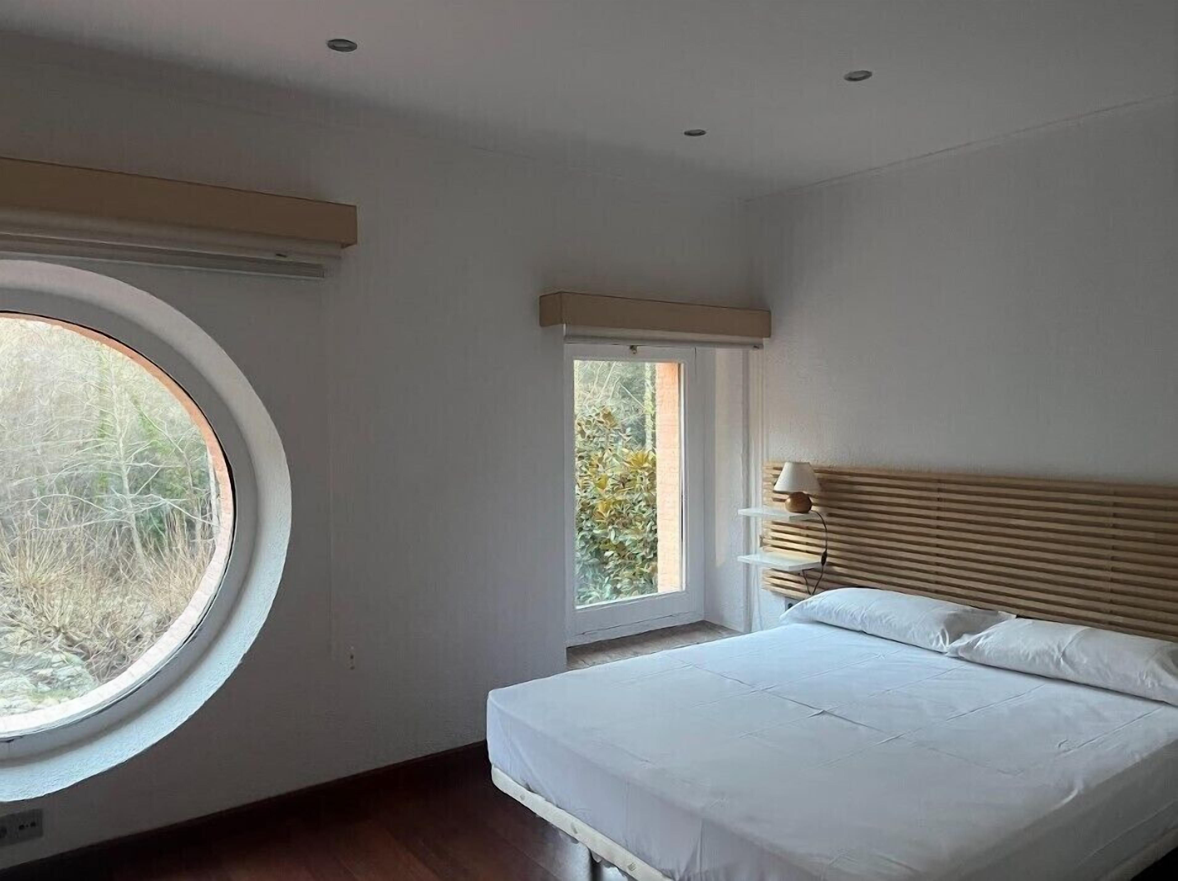 A bedroom with a round window, a bed with white bedding, a bedside table with a lamp, and a window with a view of trees.