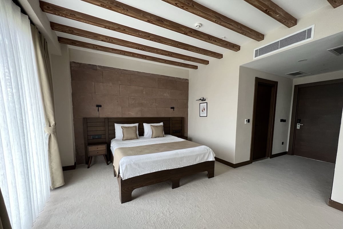 Un dormitorio con una cama grande, vigas de madera en el techo, una pared de acento de piedra y una puerta a la derecha.