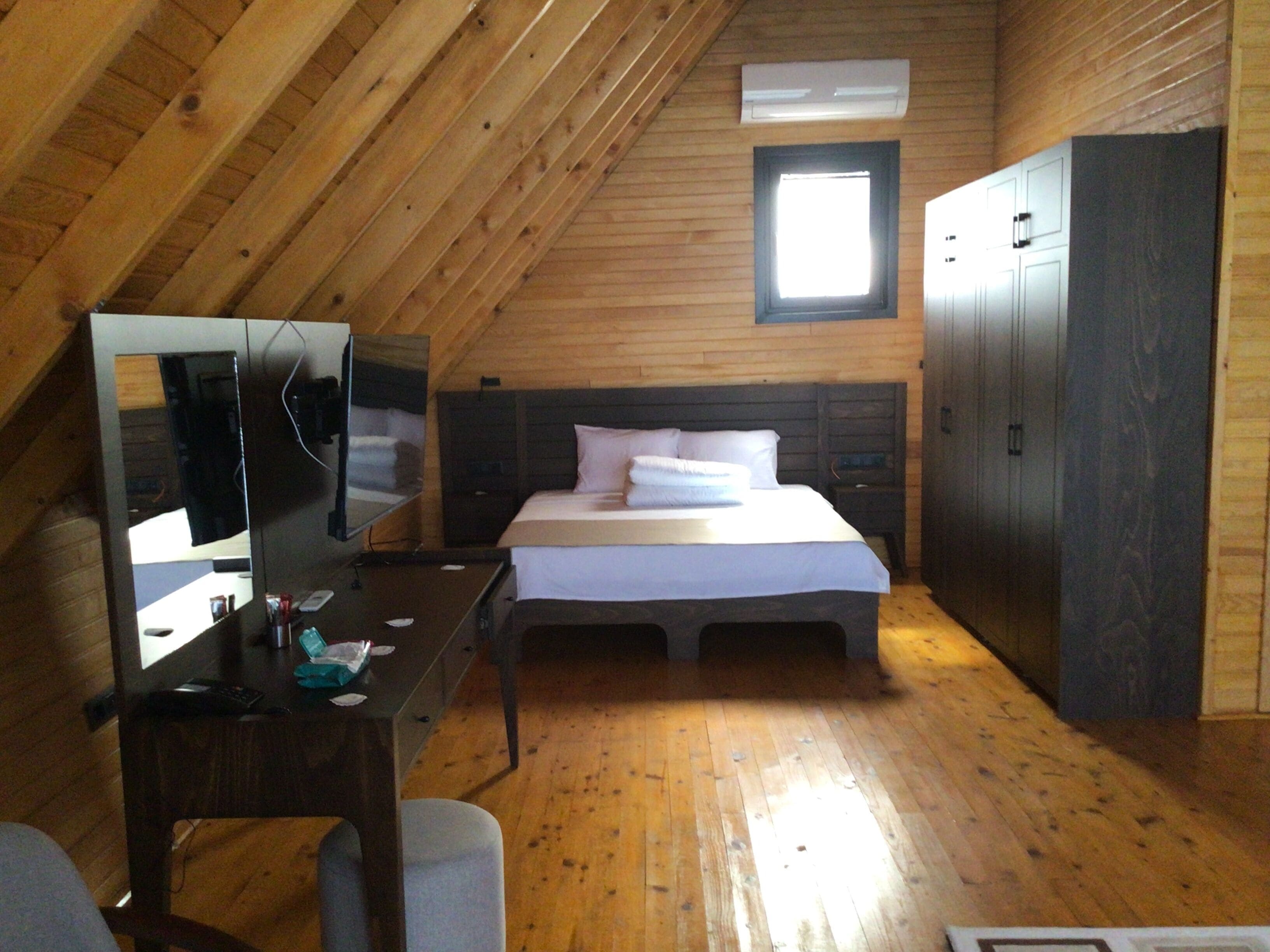 Ein Zimmer mit schrägverdeckter Decke, Holzwänden, einem Bett mit Kissen, einem Nachttisch mit Spiegel, einem Fernseher und einem Kleiderschrank.