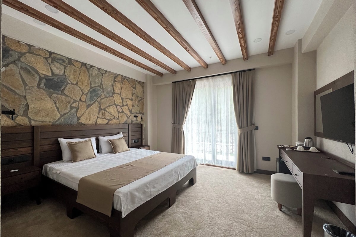 Un dormitorio con pared de piedra, techo de madera, cama con cabecera, escritorio, silla y televisor.