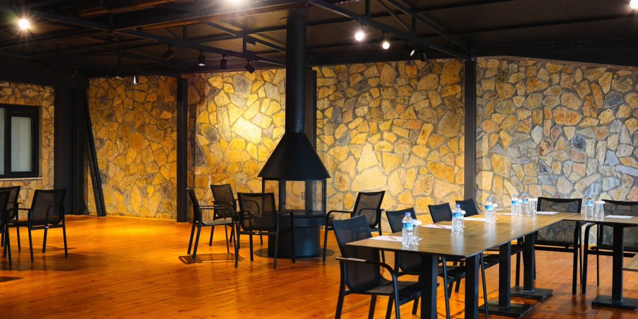 Un salón de conferencias con pared de piedra, chimenea negra, mesas y sillas de madera.