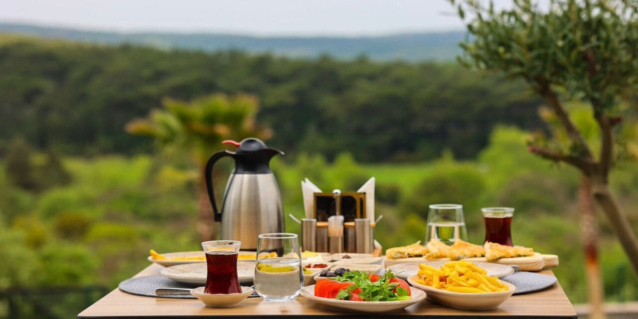 Een gedekte tafel met diverse gerechten, een koffiepot en glazen water en thee, met een natuurlandschap op de achtergrond.