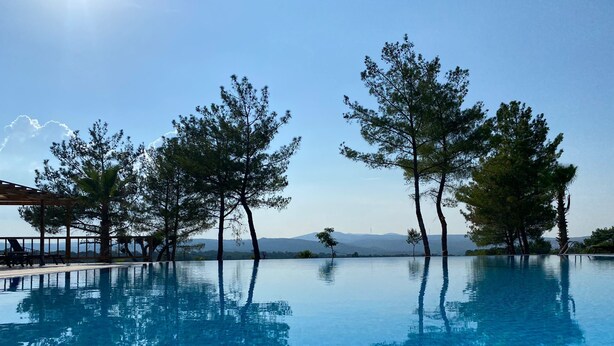 Uma piscina infinita azul cristalina com reflexos de árvores e montanhas ao fundo.