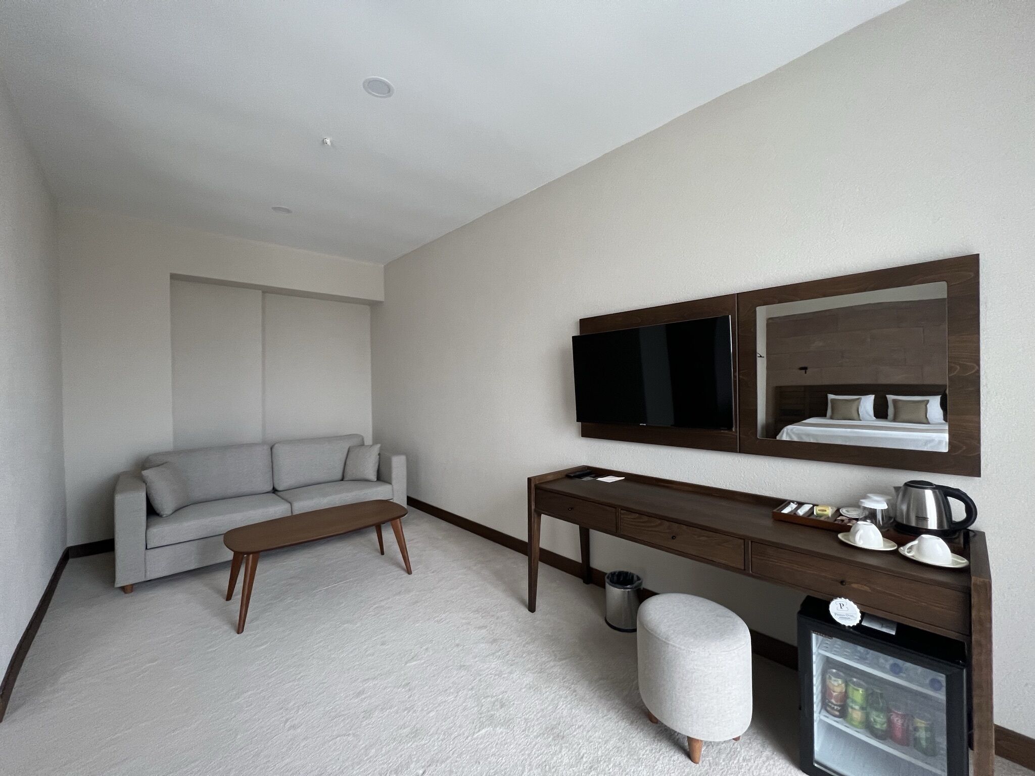 Une chambre d’hôtel moderne avec un canapé gris, un bureau en bois, une télévision, un miroir et un mini-bar.