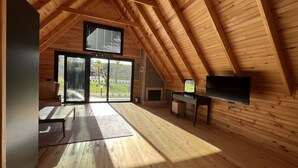 Um quarto espaçoso no sótão com vigas de madeira, uma grande porta de vidro deslizante, uma TV de tela plana e uma pequena escrivaninha.
