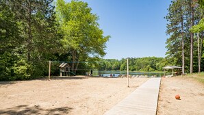 A sandy beach with a wooden walkway leading to a lake, surrounded by trees and a pavilion.