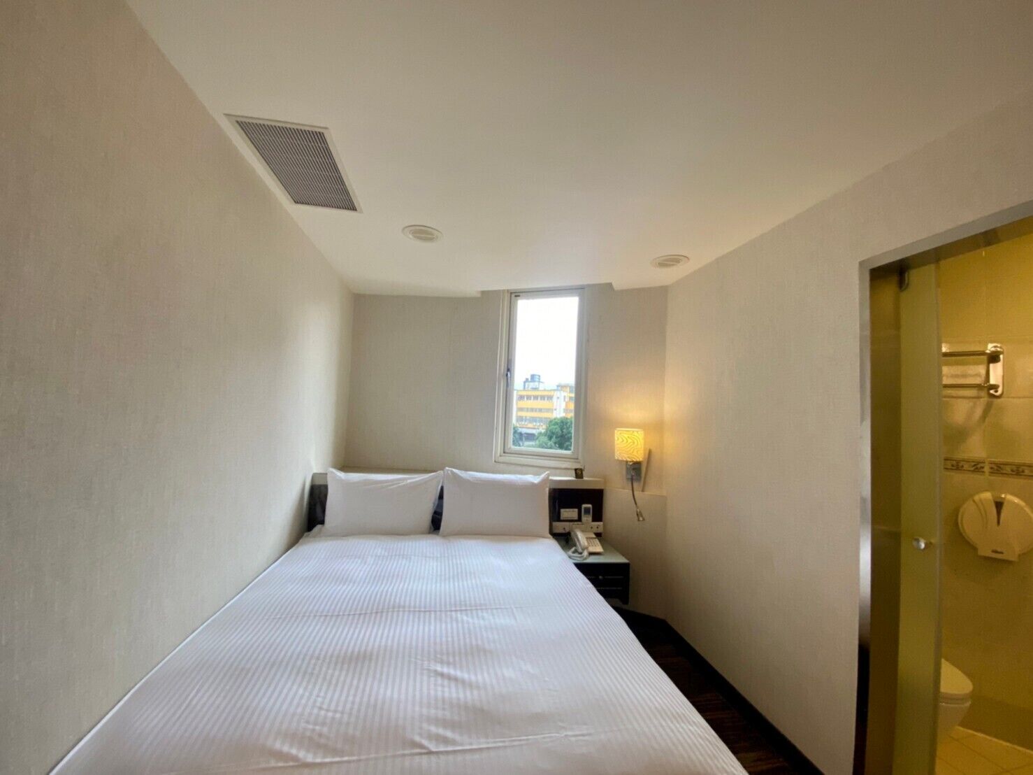 Une petite chambre d’hôtel étroite, avec un lit simple, une table de chevet avec une lampe, une fenêtre et une porte donnant sur une salle de bain.