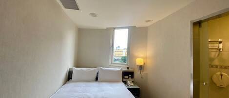 Une petite chambre d’hôtel étroite, avec un lit simple, une table de chevet avec une lampe, une fenêtre et une porte donnant sur une salle de bain.