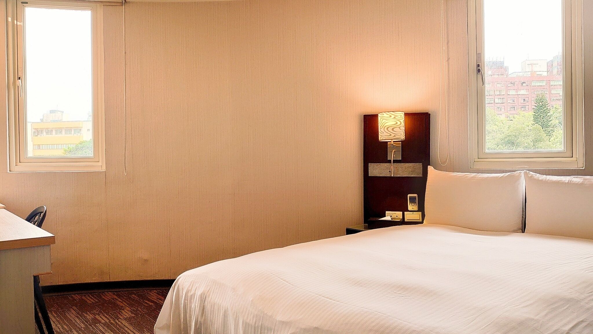 Ein Hotelzimmer mit Bett, Nachttisch, Lampe und Blick auf Gebäude durch das Fenster.