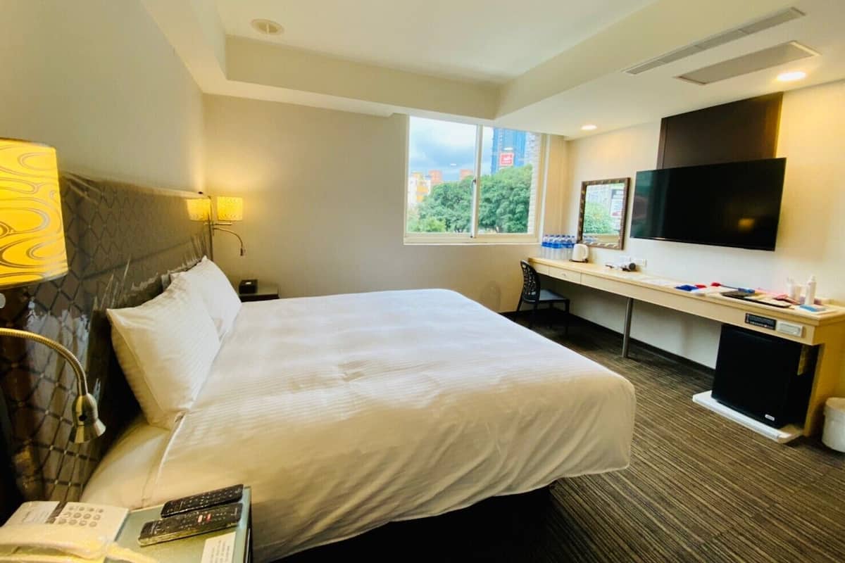Une chambre d’hôtel avec un grand lit, un bureau, une télévision et une fenêtre donnant sur des immeubles et des arbres.