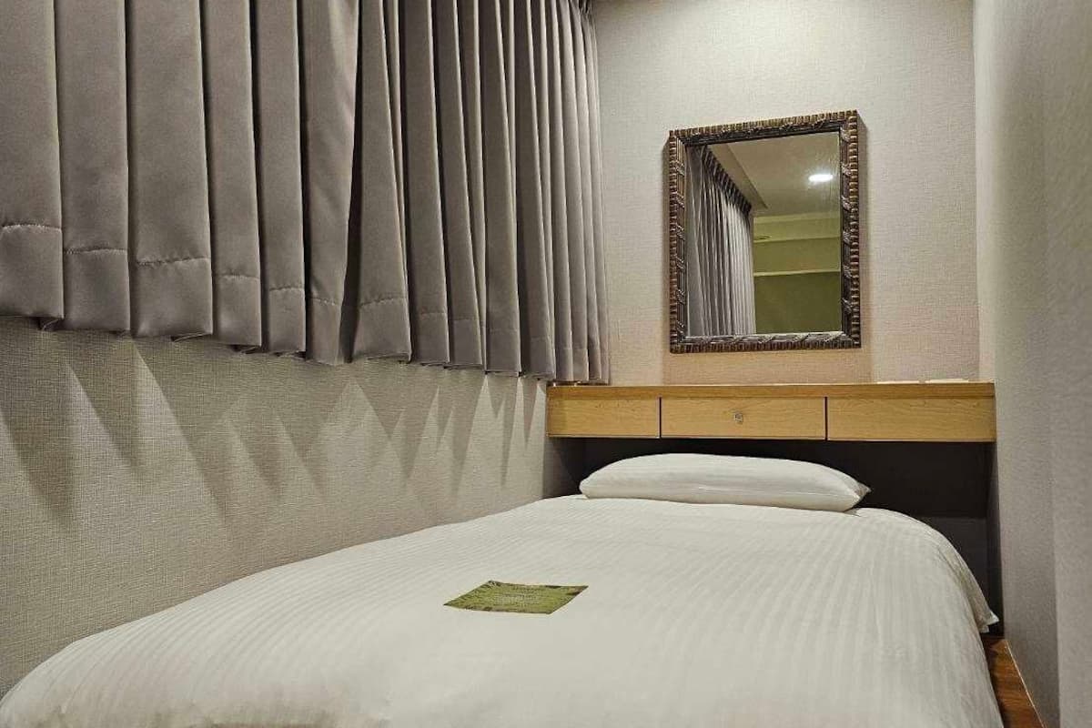 Un lit bien fait, avec une couette blanche et un couvre-lit vert, une tête de lit en bois, un miroir avec un cadre décoratif et des rideaux gris.