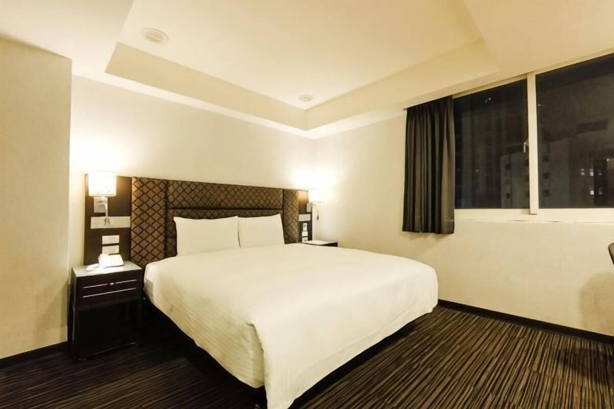 Une chambre d’hôtel avec un grand lit, deux tables de chevet avec des lampes, une fenêtre avec des rideaux et un sol recouvert de moquette.