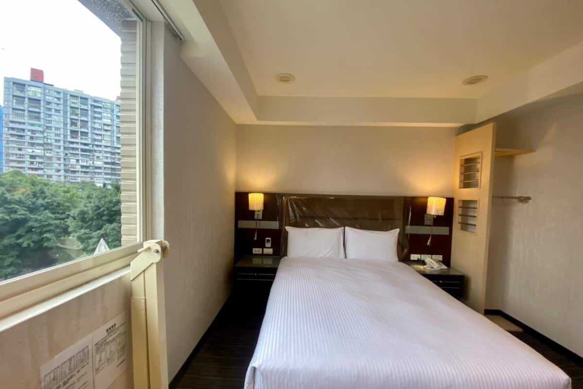 Une chambre d’hôtel avec un grand lit, deux tables de chevet avec des lampes, une fenêtre donnant sur des immeubles et des arbres, et une porte avec un porte-serviettes.