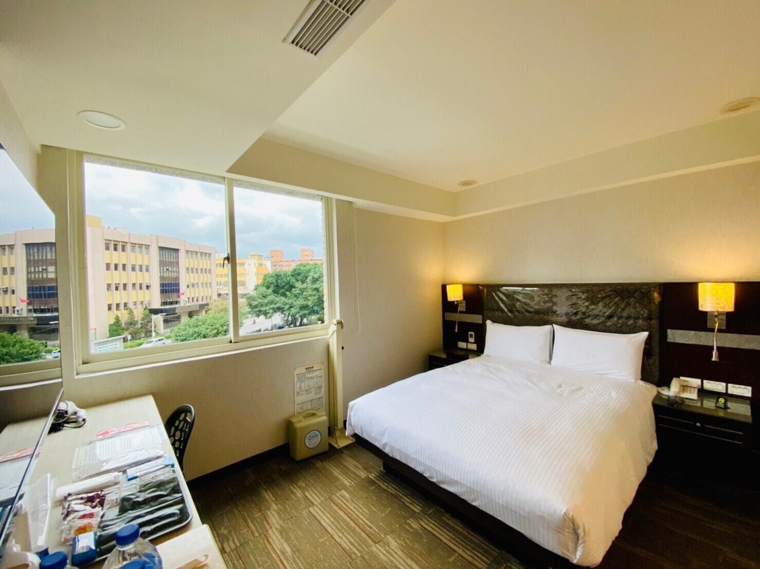Ein Hotelzimmer mit einem großen Bett, zwei Nachttischen mit Lampen, einem Schreibtisch mit Stuhl, einem Fenster mit Blick auf Gebäude und Bäume sowie einer kleinen Mülleimer.