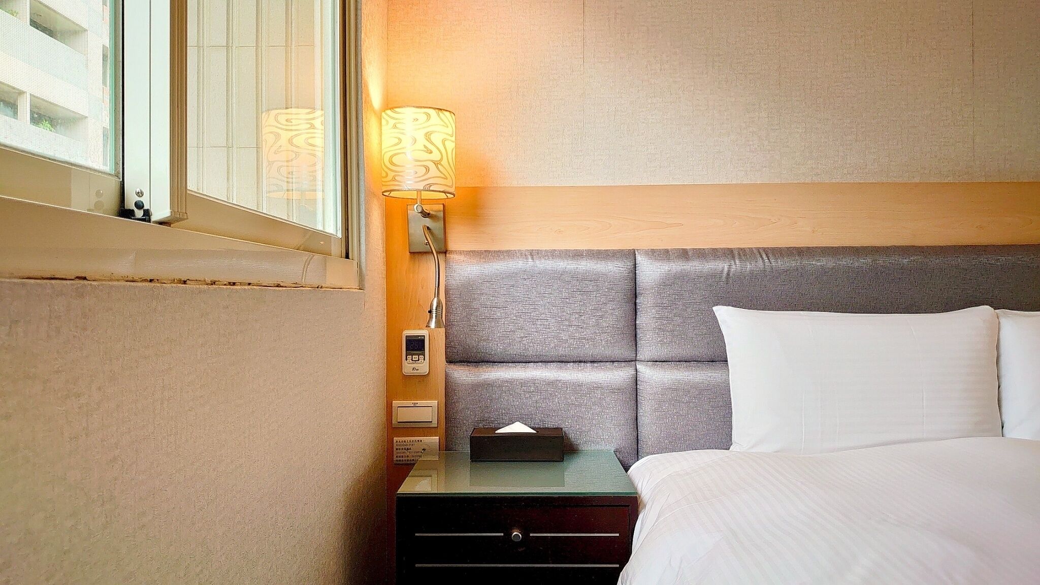 A hotel room with a bed, a nightstand, a lamp, and a window.