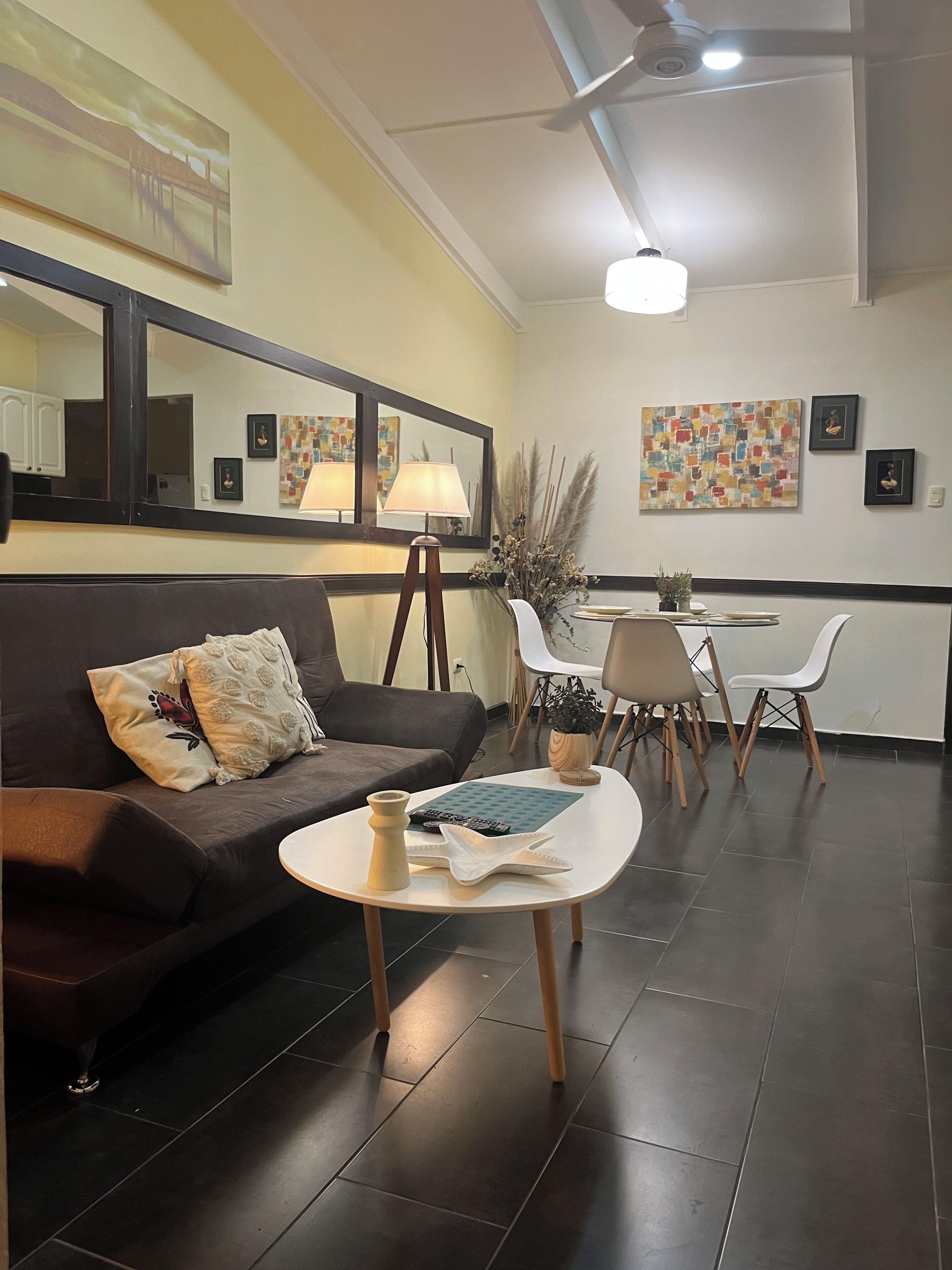 A modern living room with a dark brown sofa, a small white table, and a dining area with chairs and a lamp.