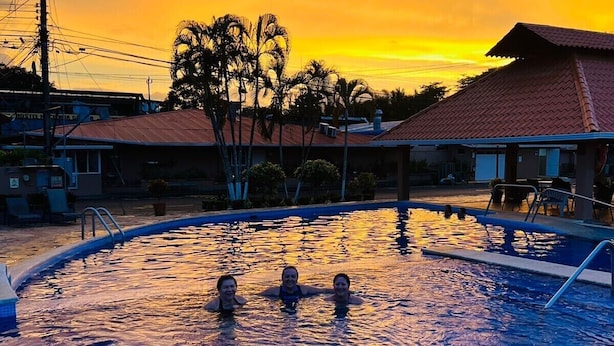 A swimming pool with three people in the water, surrounded by buildings and palm trees, during sunset.