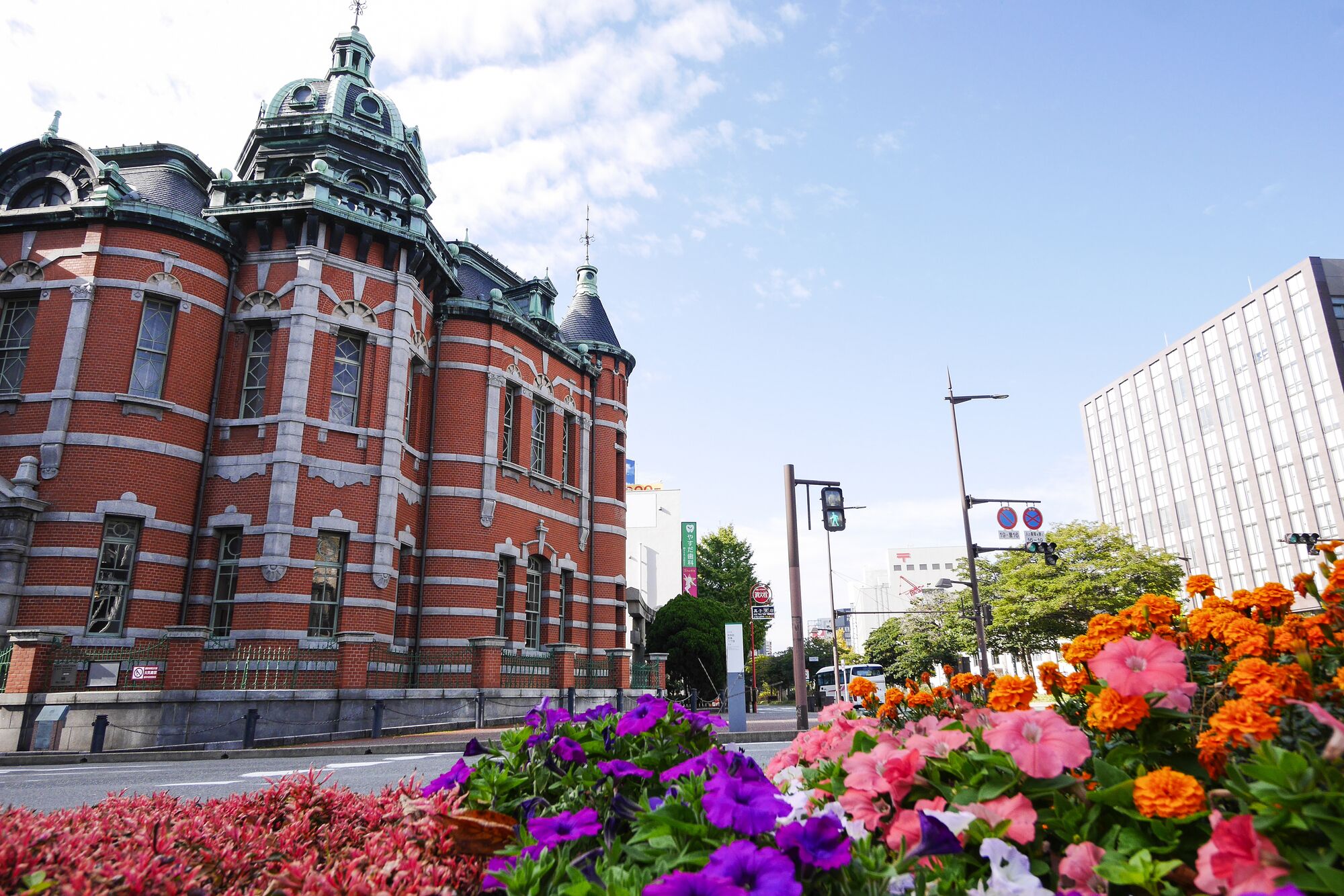A historic building with distinctive architecture, a street with traffic lights, and a flower bed in the foreground.