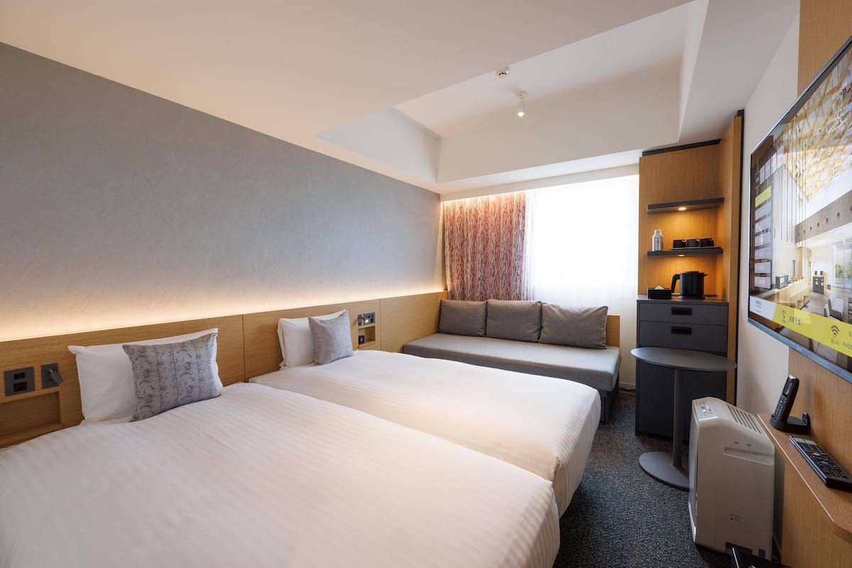 Une chambre d’hôtel avec deux lits, un coin salon, un bureau avec un ordinateur et une grande fenêtre avec des rideaux.