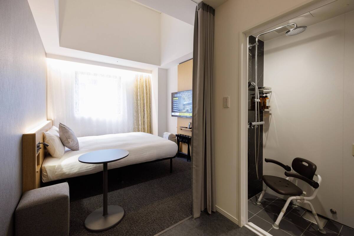 Une chambre d’hôtel avec un lit, une petite table, une chaise, un espace douche et un téléviseur fixé au mur.
