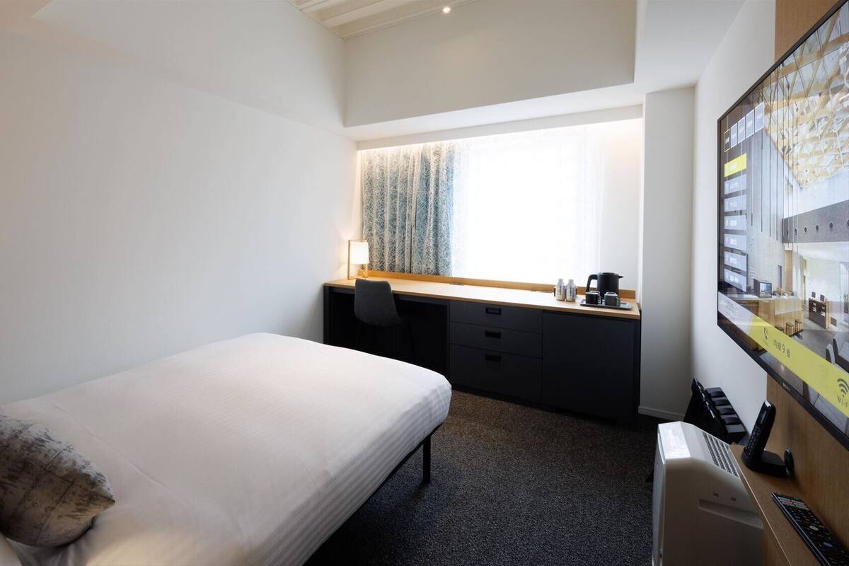 Une chambre d’hôtel avec un lit simple, un bureau avec un ordinateur, une petite kitchenette et une grande fenêtre avec des rideaux à motifs.