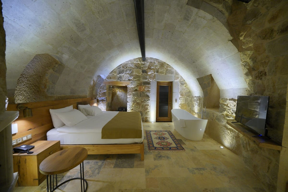 A cozy bedroom with a stone arch ceiling, a bed, a desk, a chair, a bathtub, and a television.