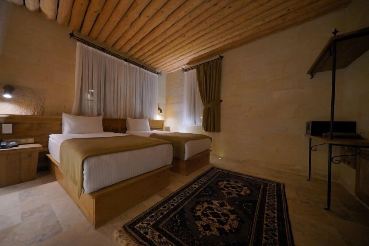 A cozy bedroom with a wooden ceiling, two beds, a nightstand, a desk, and a patterned rug.