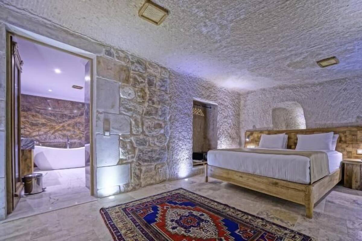 A bedroom with a stone wall, a wooden bed, a patterned rug, and a bathroom with a bathtub.