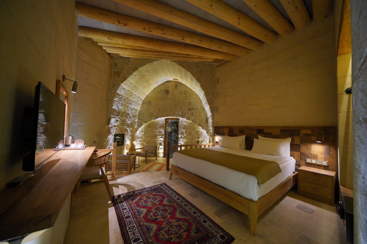 A cozy bedroom with a wooden bed, a desk with a chair, and a stone archway in the background.