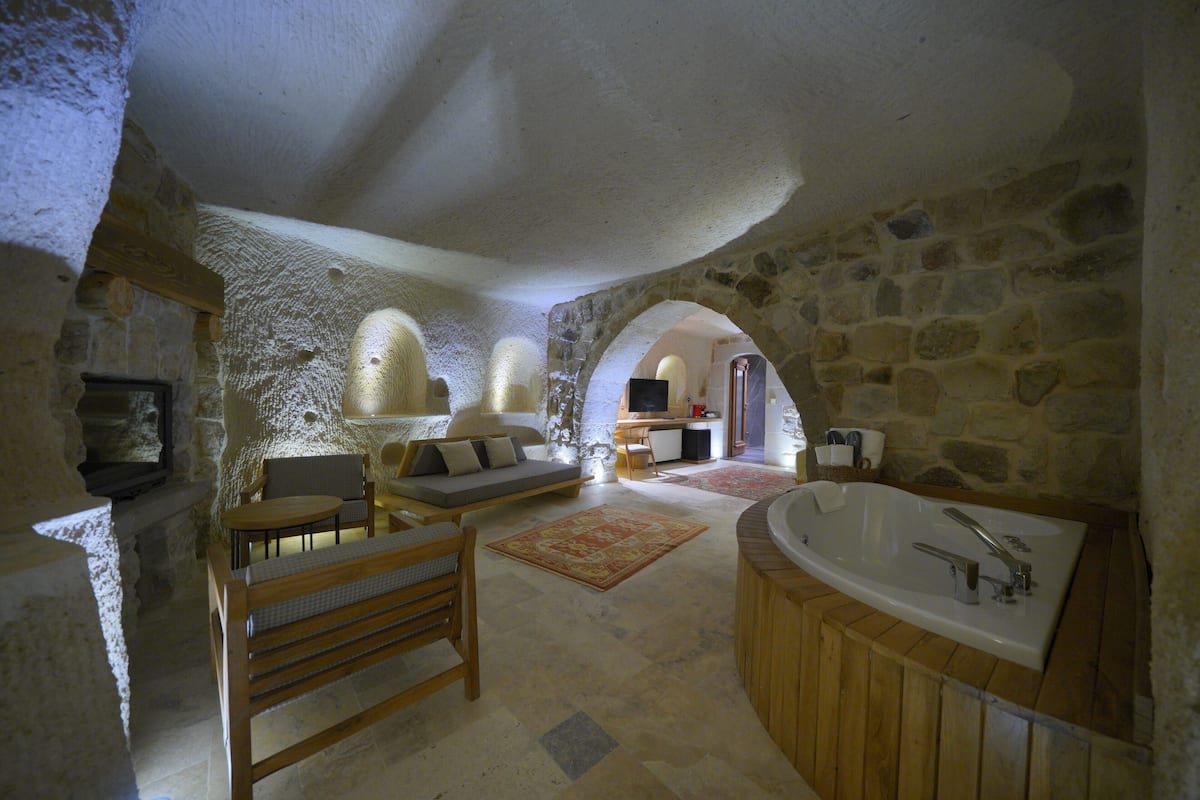 A stone-walled room with a wooden bench, a sofa, a round table, and a bathtub.
